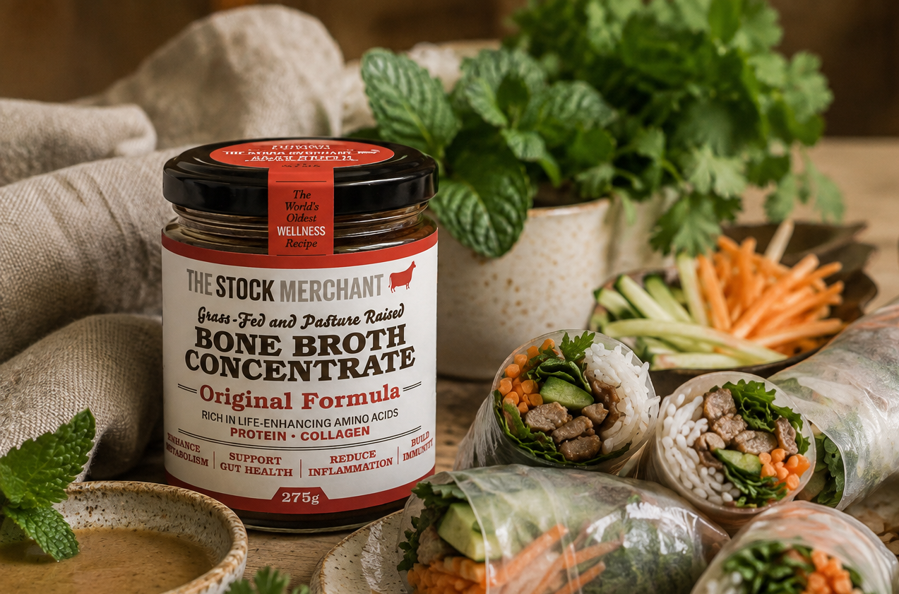 A jar of The Stock Merchant Bone Broth Concentrate stands on a rustic table beside spring rolls filled with vegetables and meat, with herbs and bowls of sauce in a styled food setting.