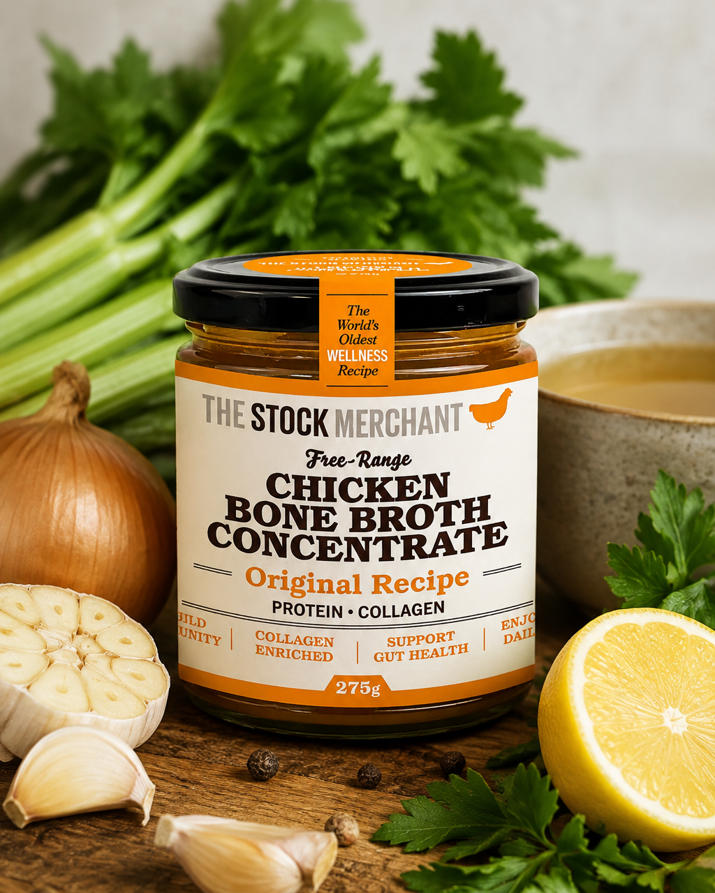 A jar of The Stock Merchant Free-Range Chicken Bone Broth Concentrate sits on a rustic wooden table surrounded by fresh ingredients including celery, garlic, lemon, parsley, and a bowl of broth.